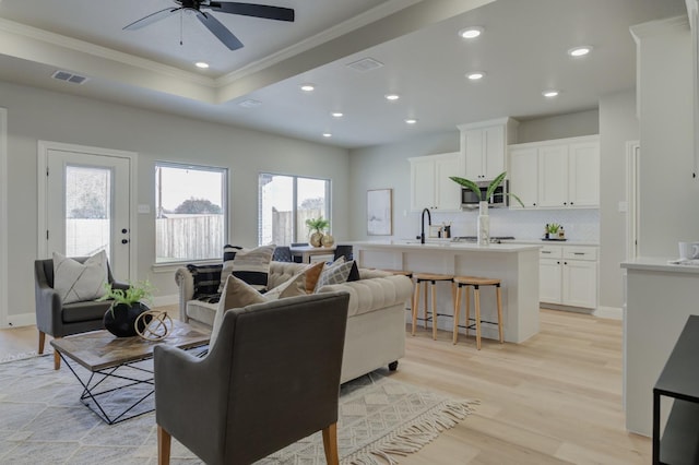 living room featuring sink, crown molding, light hardwood / wood-style flooring, a tray ceiling, and ceiling fan