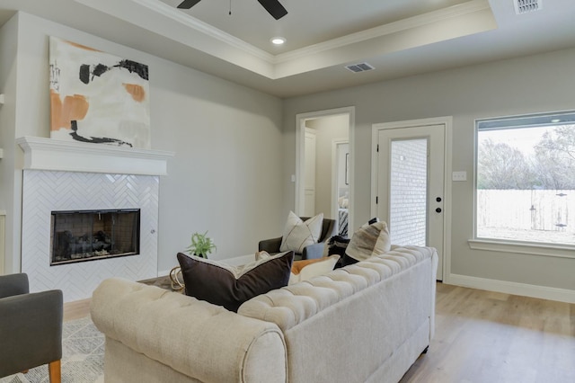 living room with a tiled fireplace, crown molding, a raised ceiling, and light hardwood / wood-style flooring