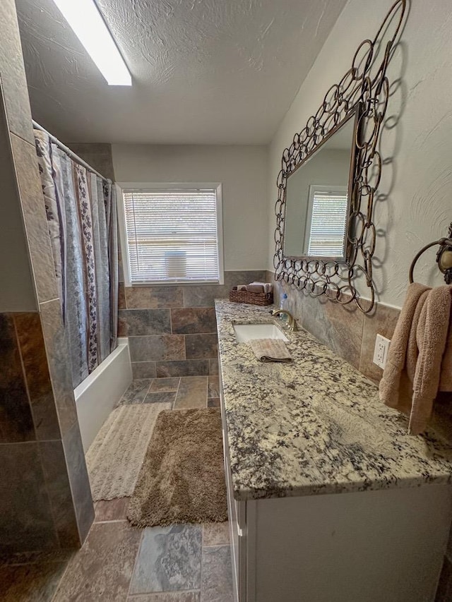 full bathroom with a textured ceiling, vanity, tile walls, stone finish floor, and shower / bathtub combination with curtain