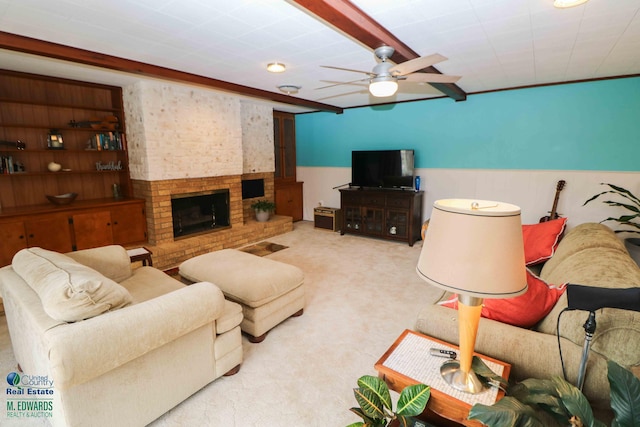 living room with beam ceiling, light colored carpet, a ceiling fan, a brick fireplace, and wainscoting