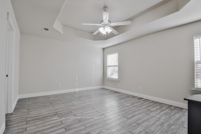 empty room with a raised ceiling, ceiling fan, a healthy amount of sunlight, and light wood-type flooring