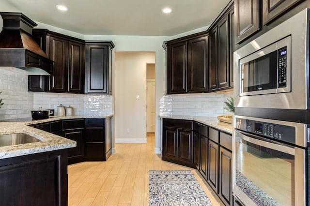 kitchen with tasteful backsplash, custom exhaust hood, light stone counters, light hardwood / wood-style floors, and stainless steel appliances