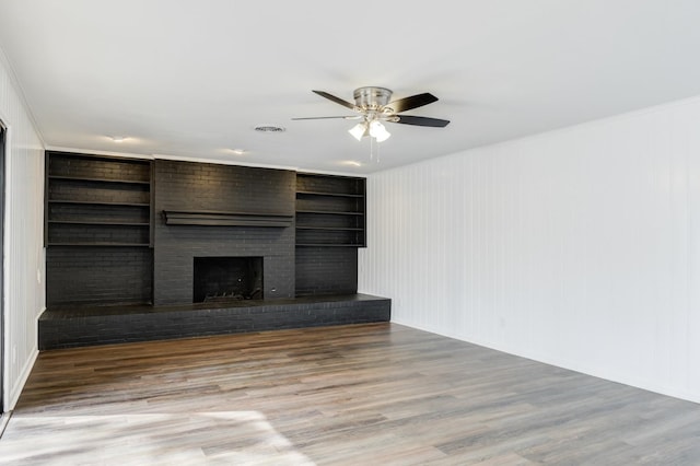 unfurnished living room featuring ceiling fan, built in features, wood-type flooring, and a brick fireplace