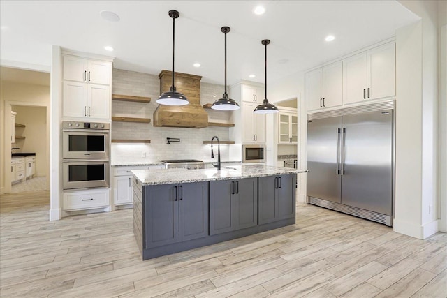 kitchen with white cabinetry, light stone counters, built in appliances, pendant lighting, and a kitchen island with sink