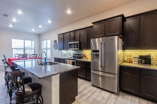 kitchen with sink, appliances with stainless steel finishes, a center island with sink, light stone countertops, and a kitchen bar