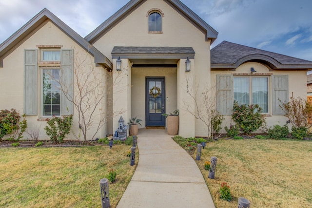 doorway to property featuring stucco siding, a yard, and roof with shingles
