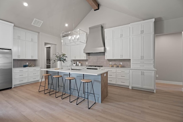 kitchen featuring custom range hood, a center island with sink, white cabinets, and stainless steel refrigerator