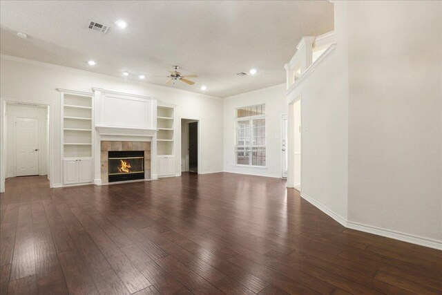 unfurnished living room with crown molding, built in features, ceiling fan, a tiled fireplace, and dark hardwood / wood-style flooring