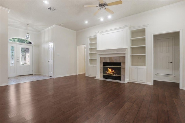 unfurnished living room featuring a tile fireplace, ceiling fan, wood-type flooring, ornamental molding, and built in shelves