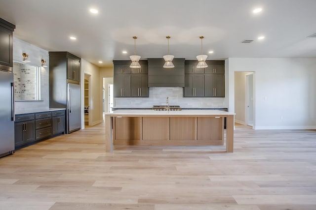 kitchen with stainless steel built in refrigerator, sink, tasteful backsplash, hanging light fixtures, and a kitchen island with sink