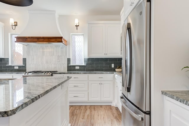 kitchen featuring custom exhaust hood, white cabinetry, stainless steel fridge, stone counters, and decorative backsplash