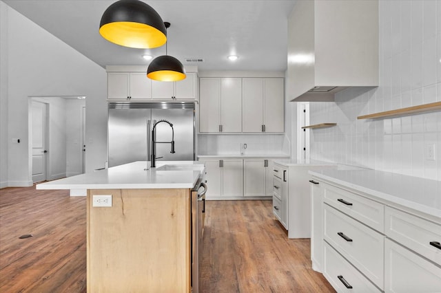 kitchen featuring pendant lighting, white cabinets, a center island with sink, built in fridge, and wall chimney exhaust hood