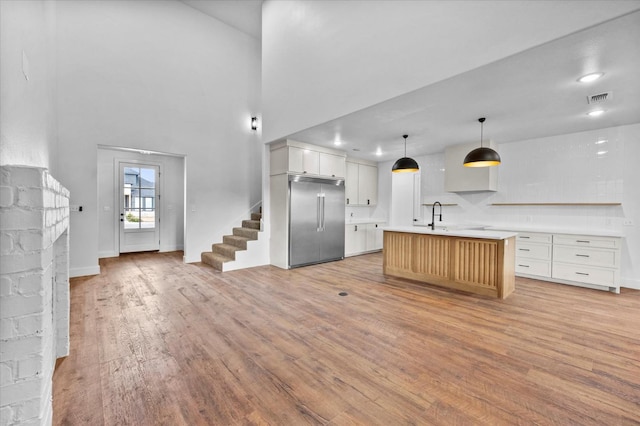 kitchen with decorative light fixtures, a kitchen island with sink, built in fridge, light hardwood / wood-style floors, and white cabinets