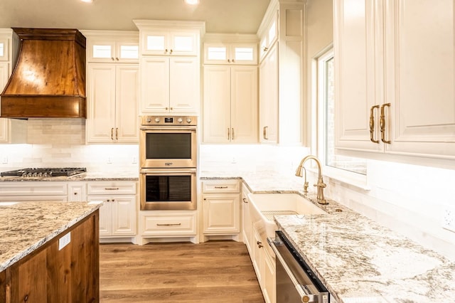 kitchen featuring sink, hardwood / wood-style floors, stainless steel appliances, light stone countertops, and custom range hood