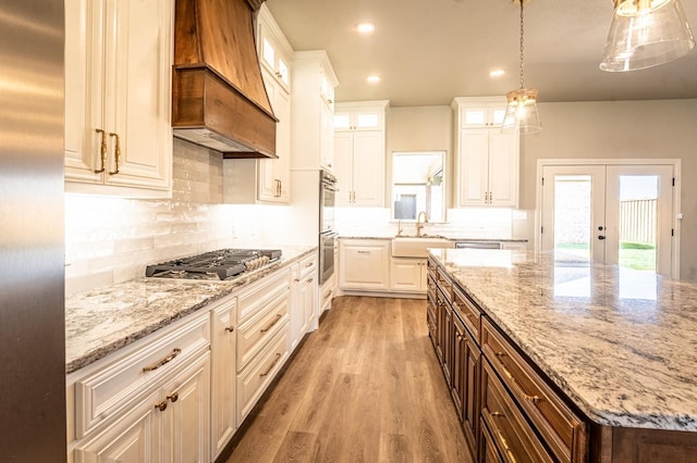 kitchen featuring appliances with stainless steel finishes, light stone countertops, custom range hood, white cabinets, and decorative light fixtures