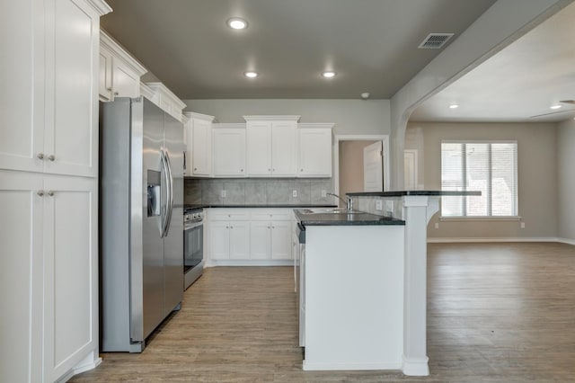 kitchen featuring appliances with stainless steel finishes, white cabinets, a center island with sink, dark stone counters, and light wood-type flooring