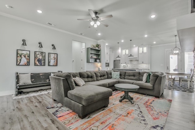 living room with crown molding, sink, ceiling fan with notable chandelier, and light wood-type flooring