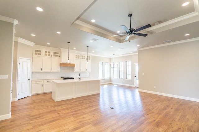 kitchen featuring white cabinets, backsplash, hanging light fixtures, a kitchen island with sink, and a raised ceiling