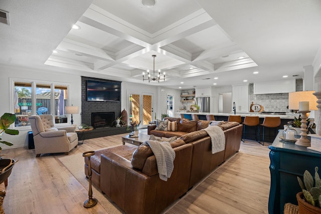 living room with light wood-type flooring, a chandelier, coffered ceiling, a brick fireplace, and beam ceiling