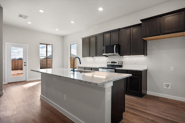 kitchen featuring sink, appliances with stainless steel finishes, dark hardwood / wood-style floors, light stone counters, and an island with sink