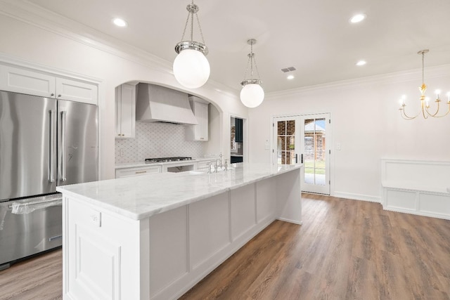 kitchen featuring decorative light fixtures, wall chimney range hood, an island with sink, stainless steel appliances, and white cabinets