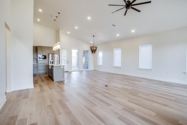 unfurnished living room with high vaulted ceiling, ceiling fan with notable chandelier, and light hardwood / wood-style floors