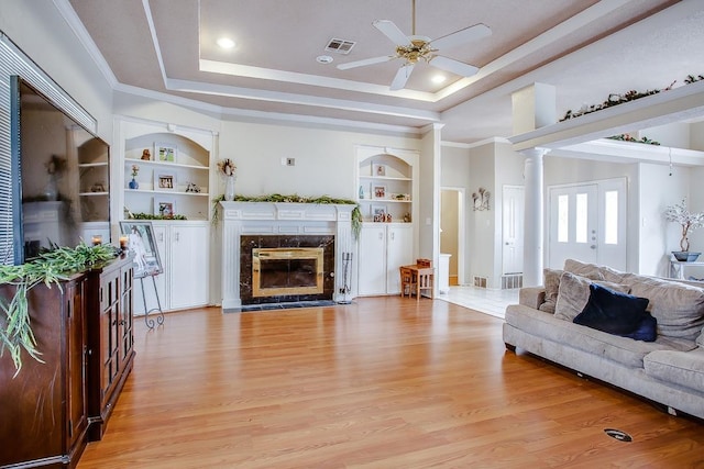 living room featuring built in shelves, light wood-type flooring, a raised ceiling, a fireplace, and decorative columns