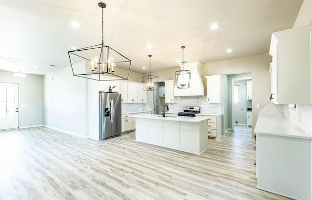 kitchen with premium range hood, white cabinetry, stainless steel appliances, an island with sink, and decorative light fixtures