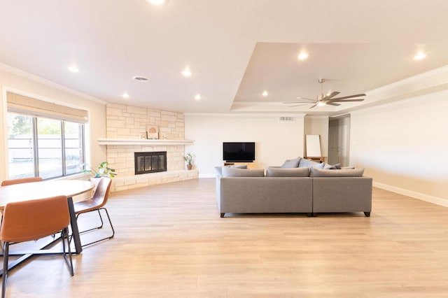 living room featuring a stone fireplace, ornamental molding, a tray ceiling, ceiling fan, and light hardwood / wood-style floors