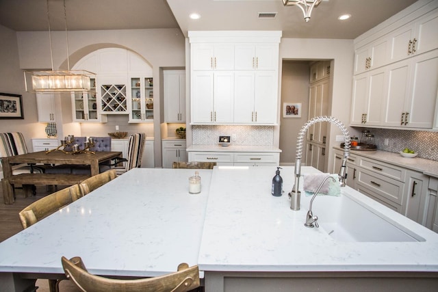 kitchen with light stone counters, a sink, an island with sink, glass insert cabinets, and pendant lighting