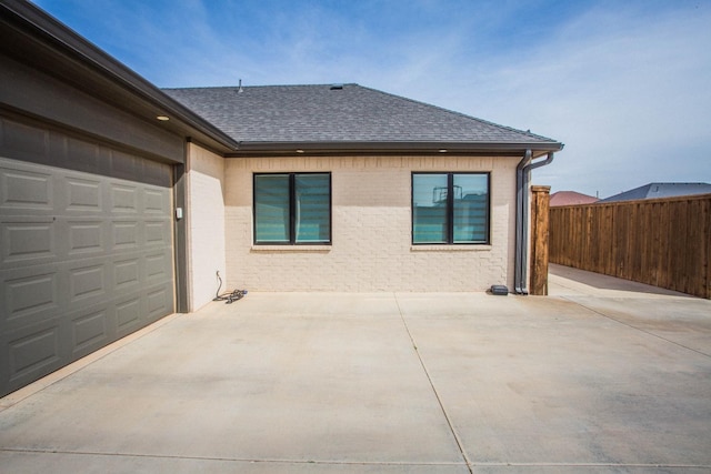 exterior space featuring driveway, roof with shingles, an attached garage, fence, and brick siding