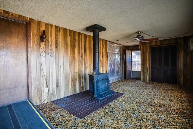 unfurnished living room with wooden walls, carpet floors, a wood stove, ceiling fan, and a textured ceiling