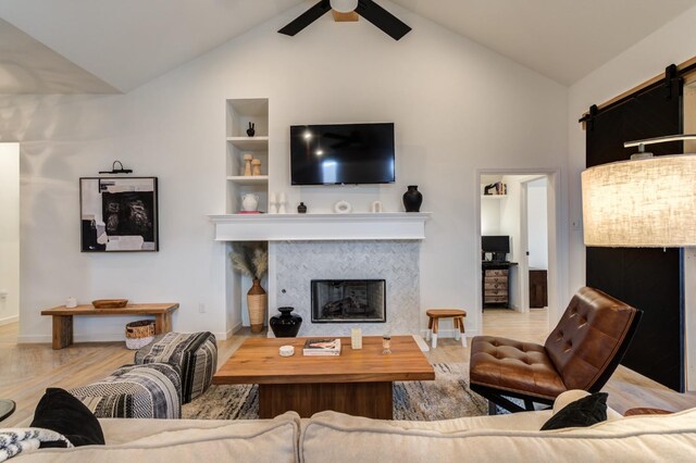 living room featuring light hardwood / wood-style flooring, ceiling fan, high vaulted ceiling, built in shelves, and a barn door