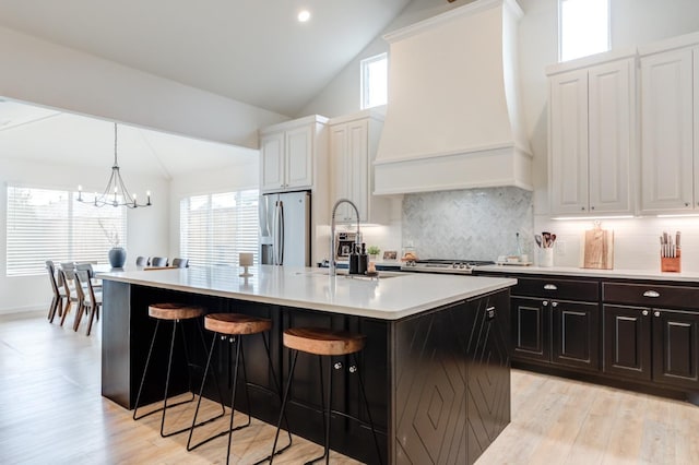 kitchen featuring sink, custom exhaust hood, stainless steel fridge with ice dispenser, a kitchen island with sink, and white cabinets