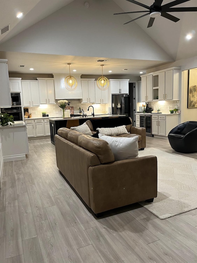 living room with ceiling fan, beverage cooler, high vaulted ceiling, and light hardwood / wood-style flooring