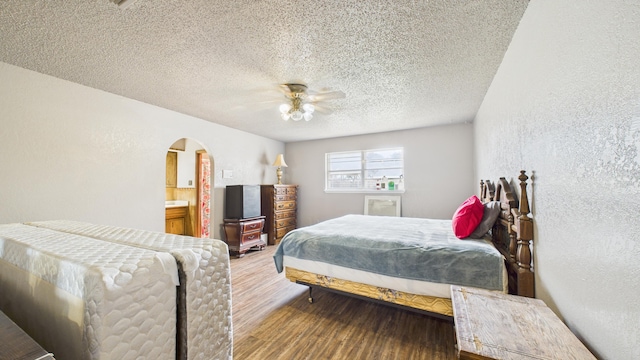 bedroom featuring ceiling fan, a textured wall, a textured ceiling, arched walkways, and wood finished floors