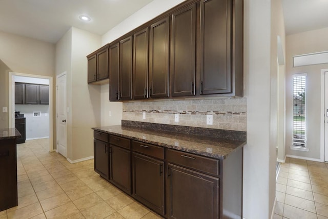 kitchen with light tile patterned floors, decorative backsplash, dark brown cabinetry, dark stone countertops, and baseboards