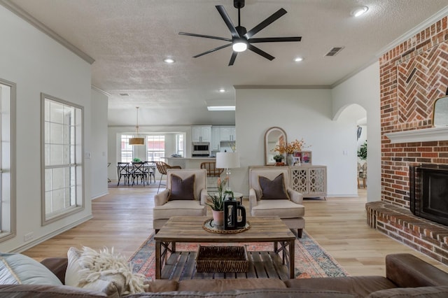 living room with ceiling fan, ornamental molding, a textured ceiling, a brick fireplace, and light wood-type flooring