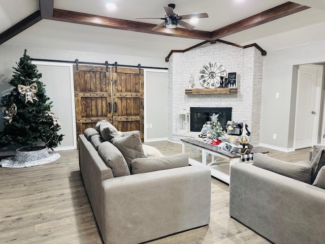 living room with ceiling fan, a fireplace, light hardwood / wood-style floors, a barn door, and beamed ceiling