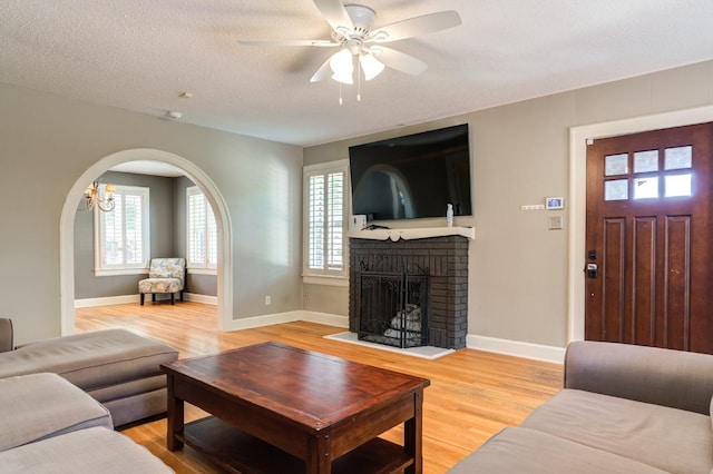 living room with a brick fireplace, light hardwood / wood-style flooring, a textured ceiling, and ceiling fan