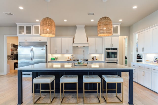 kitchen featuring premium range hood, appliances with stainless steel finishes, pendant lighting, a kitchen island with sink, and backsplash