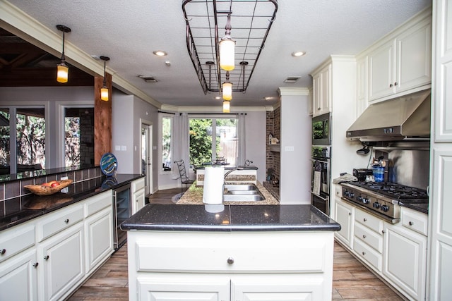 kitchen with an island with sink, beverage cooler, hanging light fixtures, stainless steel appliances, and light hardwood / wood-style flooring