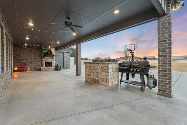 patio terrace at dusk featuring ceiling fan, area for grilling, and an outdoor stone fireplace