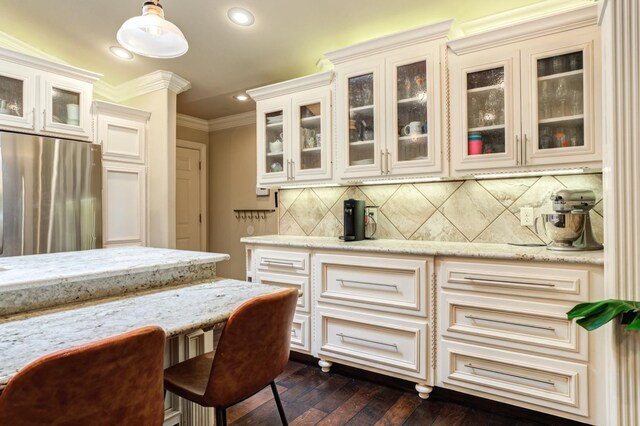 kitchen with tasteful backsplash, hanging light fixtures, stainless steel refrigerator, ornamental molding, and light stone countertops