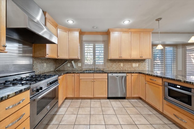 kitchen with wall chimney range hood, sink, appliances with stainless steel finishes, hanging light fixtures, and dark stone counters