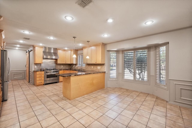 kitchen with light brown cabinetry, decorative light fixtures, kitchen peninsula, stainless steel appliances, and wall chimney exhaust hood