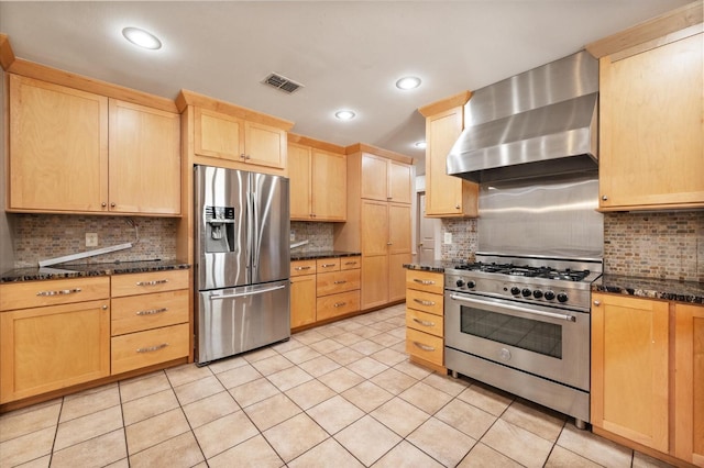 kitchen with appliances with stainless steel finishes, wall chimney range hood, light brown cabinetry, and dark stone counters