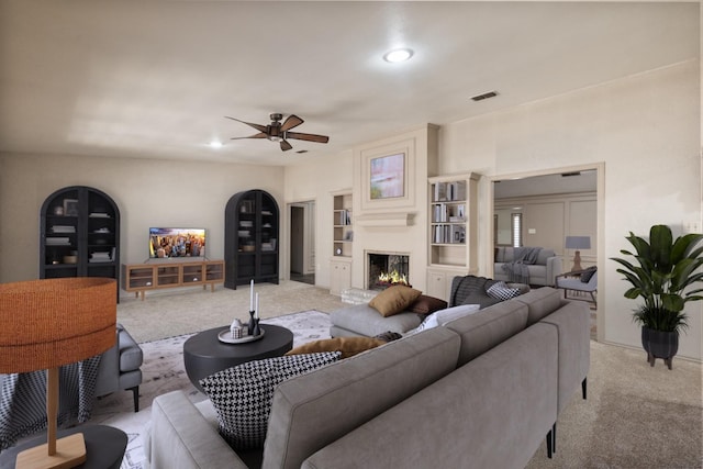 living room with built in features, light colored carpet, a large fireplace, and ceiling fan