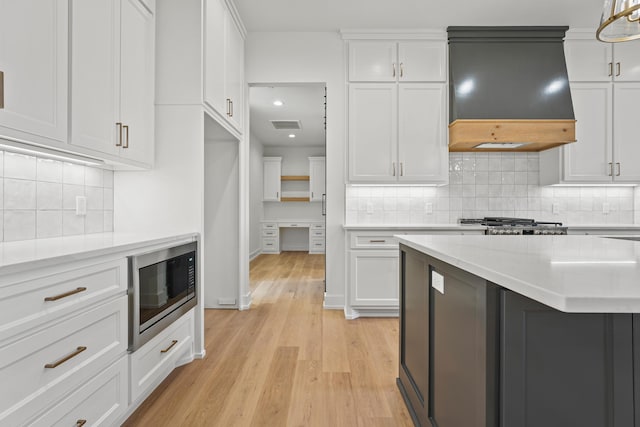 kitchen with decorative light fixtures, built in microwave, white cabinetry, custom exhaust hood, and light hardwood / wood-style flooring