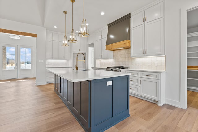 kitchen with custom exhaust hood, range, hanging light fixtures, an island with sink, and white cabinets